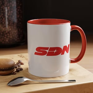 May include: A white ceramic mug with a red interior and handle. The mug displays the red letters "SDN" with a star design in the center. A spoon and a macaron are on a wooden surface.