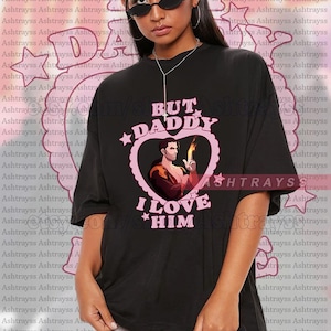 May include: Black unisex t-shirt with a pink heart graphic featuring a cartoon image and the text "BUT DADDY I LOVE HIM". The shirt also has the word "ASHTRAYS" printed on it.