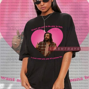 May include: Black unisex t-shirt with the text "Trust me gay is in, gay is hot" and "I want some gay, gay it's gonna be." The shirt features a heart graphic with a man giving a thumbs up.