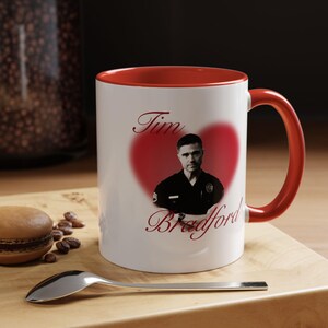 Pode incluir: Uma caneca de cerâmica branca com interior e alça vermelhos. A frente apresenta um retrato em preto e branco de um homem com uniforme de polícia centralizado dentro de um coração vermelho, com o texto Tim Bradford em fonte cursiva.