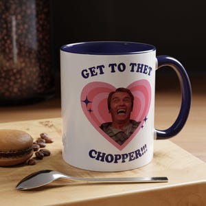 Könnte beinhalten: Weißer Keramikbecher mit marineblauem Innenraum und Griff. Der Becher zeigt den Text "GET TO THET CHOPPER!!!" und ein rosa Herz mit einem Bild eines Mannes. Ein Löffel und ein Macaron sind im Vordergrund.