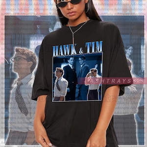 May include: Black unisex t-shirt featuring the text "HAWK & TIM" above a framed image of three men. The shirt is worn by a person wearing sunglasses. The background has a repeating pattern of text.