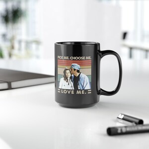 May include: A black ceramic mug featuring a graphic print of two medical professionals in scrubs. The text above the image reads PICK ME. CHOOSE ME. and the text below reads LOVE ME. The background of the graphic consists of horizontal stripes.