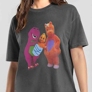 May include: A charcoal grey short-sleeve t-shirt featuring a graphic print of three characters. On the left, a purple dinosaur with a green belly. In the centre, a small orange creature wrapped in a blue blanket. On the right, an orange unicorn.