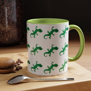 Puede incluir: Taza de cerámica blanca con interior y asa verdes. La taza está decorada con un patrón repetido de lagartos verdes de dibujos animados. Una cuchara y un macaron están sobre una superficie de madera junto a la taza.