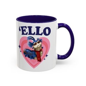 May include: White ceramic mug with a blue interior and handle. The mug features the word "'ELLO" above a cartoon worm character inside a pink heart. The worm has blue fur, a red scarf, and is set against a white background.