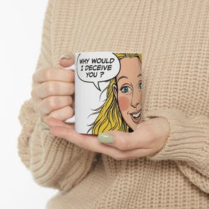 May include: A white ceramic mug featuring a pop art style illustration of a blonde person with blue eyes. A speech bubble above the person contains the black text, "WHY WOULD I DECEIVE YOU ?". The mug has a smooth finish and a standard cylindrical shape.