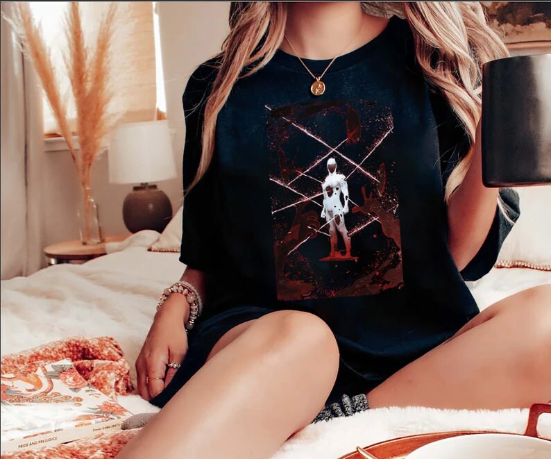 May include: Black t-shirt featuring a graphic design of a figure in a white suit with red accents and lines. The design is centered on the shirt. The shirt is worn by a person.