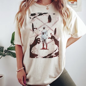 May include: Cream-colored t-shirt featuring a graphic design. The design includes a white figure with black spots, surrounded by red splatters and dark silhouettes of hands. The shirt is a relaxed fit.