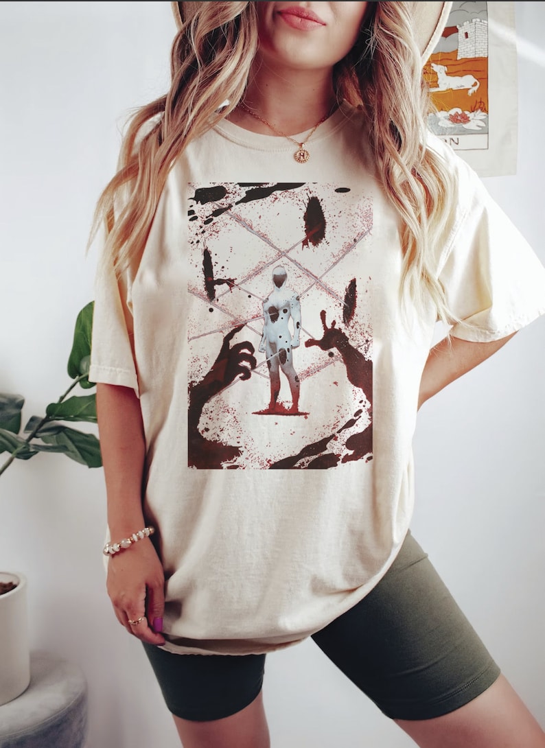 May include: Cream-colored t-shirt featuring a graphic design. The design includes a white figure with black spots, surrounded by red splatters and dark silhouettes of hands. The shirt is a relaxed fit.
