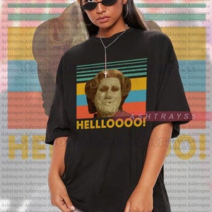 May include: Black oversized t-shirt featuring a graphic of a person with a face mask, set against a retro striped background. The shirt has the word "HELLLOOOO!" in yellow. The model is wearing a 2XL shirt.