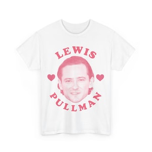 Puede incluir: Camiseta blanca con un gráfico rosa de la cara de Lewis Pullman rodeado del texto "LEWIS PULLMAN" y dos corazones rosas.