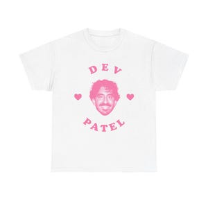 May include: White t-shirt with a pink graphic featuring the name "DEV PATEL" in a stylized font, with a portrait of Dev Patel in the center. Two pink hearts flank the portrait. A casual, fan-themed apparel item.