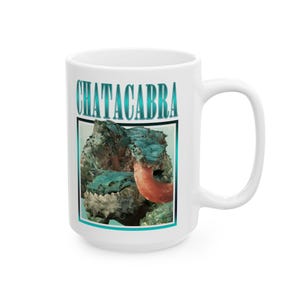 Może przedstawiać: Biały ceramiczny kubek z nadrukiem graficznym przedstawiającym teksturowane niebieskie stworzenie z długim różowym językiem. Nad stworzeniem znajduje się napis CHATACABRA wykonany metaliczną niebieską czcionką. Wzór otoczony jest cienką niebieską ramką.