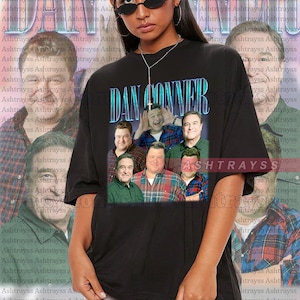 May include: A black t-shirt with a graphic print of the cast of the TV show "Dan Conner" from the show "Roseanne". The graphic is in a retro style with a blue and green color scheme. The text "Dan Conner" is printed in large, bold letters above the image of the cast.