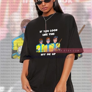 May include: Black unisex t-shirt with a cartoon design of three figures and the text "IF YOU LOOK LIKE THIS HIT ME UP" and an Etsy shop URL. The shirt is oversized and made of a soft fabric.