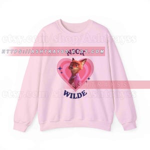 May include: A light pink sweatshirt featuring a cartoon image of Nick Wilde from Zootopia inside a pink heart. The sweatshirt has long sleeves and the name "Nick Wilde" is printed on the front.