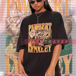 May include: Black unisex t-shirt featuring the text "PAWBERT LYNXLEY" in orange and yellow, with a cartoon image of three lynx-like characters. The shirt is oversized and made of a soft material.