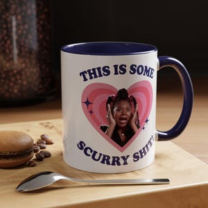 May include: White ceramic mug with a navy blue interior and handle. The mug features the text "THIS IS SOME SCURRY SHIT!" around a pink heart graphic with a woman's image. A spoon and a pastry are in the foreground.