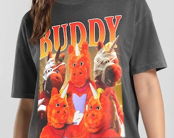 May include: A charcoal gray t-shirt featuring a graphic print of five orange plush unicorn characters. The word BUDDY is printed in large, bold, orange and yellow gradient letters above the unicorns. Two of the unicorns have white bandages.