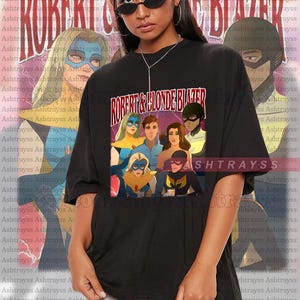 May include: Black oversized t-shirt with a superhero team graphic and the text "ROBERT & BLONDE BLAZER". This unisex shirt is designed for a relaxed fit.