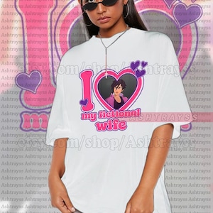以下が含まれることがあります： ピンクのハートの中に「I love my fictional wife」という文字とアニメのキャラクターの画像が入った白いユニセックスTシャツ。モデルは2XLを着用。Tシャツはジャストサイズです。