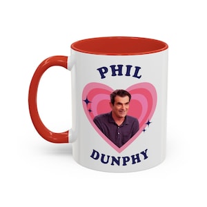 Op de afbeelding: Witte keramische mok met een rode binnenkant en handvat. De mok heeft een roze hartontwerp met de woorden "PHIL DUNPHY" in blauw. Een foto van een man staat in het hart, met blauwe steraccenten.