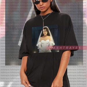 May include: Black unisex t-shirt with a graphic of a bride in a white wedding dress and veil. The shirt is oversized and has the text "ASHTRAYSS" repeated in the background. The model is wearing a 2XL.