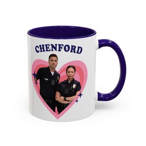 Pode incluir: Caneca de cerâmica branca com interior e alça azul marinho. A caneca apresenta a palavra "CHENFORD" em letras azul marinho acima de um gráfico de coração rosa com duas pessoas em uniformes policiais. Explosões de estrelas azuis estão no coração.