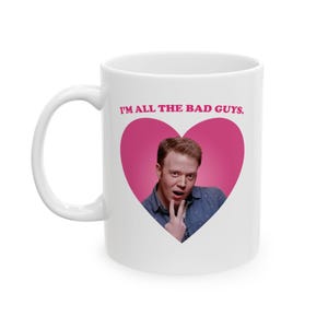 Peut inclure: Mug en céramique blanche avec l'inscription "I'M ALL THE BAD GUYS." au-dessus d'un cœur rose. À l'intérieur du cœur, un homme en chemise bleue fait le signe de la paix. La tasse a une anse blanche.