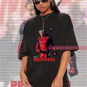 May include: Black unisex t-shirt featuring a graphic of a character in a red and black costume with a sword, and the text "THE PROPOSAL". The model is wearing a 2XL shirt. The shirt is oversized.