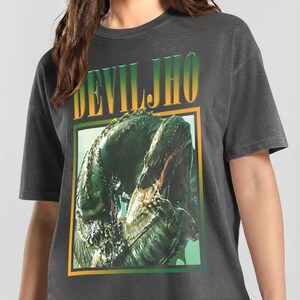 May include: A charcoal gray t-shirt featuring a graphic print of a green, reptilian creature with sharp teeth. Above the creature, the text DEVILJHO is printed in a bold, yellow-to-green gradient font. The graphic is framed by a thin yellow and green border.
