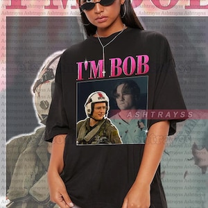 Puede incluir: Una camiseta negra con un gráfico rosa que dice "I'M BOB" y presenta dos imágenes de un hombre con uniforme de piloto. La modelo lleva una camiseta talla 2XL. Es una camiseta unisex de talla real, y para conseguir el look de talla grande, tienes que elegir +1 o +2 de tu talla original.