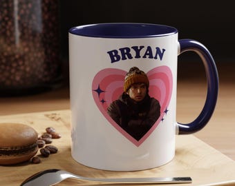 May include: A white ceramic mug with a dark blue interior and handle. The front features a pink heart graphic containing a photograph of a person wearing a beanie, with the name BRYAN printed in dark blue letters above the heart design.