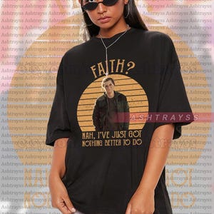 May include: Black unisex t-shirt with a graphic featuring a man's image inside a circular design, the word "FAITH?" above, and the text "NAH, I'VE JUST GOT NOTHING BETTER TO DO" below. The shirt is oversized.