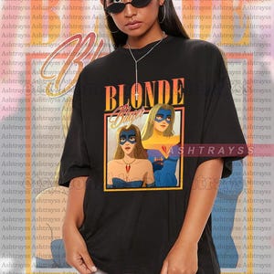 May include: Black unisex t-shirt with a graphic of two women in superhero costumes and the word "BLONDE". The shirt is oversized and has the brand name "ASHTRAYSS" printed repeatedly in the background.