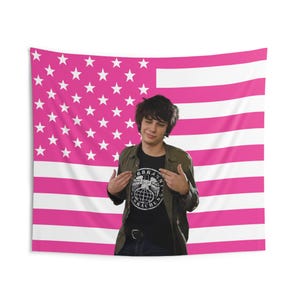 May include: A wall tapestry featuring a pink and white American flag pattern with a central graphic of a person with dark hair wearing an olive green button-down shirt over a black t-shirt with a circular logo and the text VERBRANNT BRAUCHUNG.