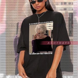 May include: Black unisex t-shirt with a graphic of Meryl Streep wearing glasses, and the text "BY ALL MEANS MOVE AT A GLACIAL PACE" and "YOU KNOW HOW THAT THRILLS ME". The shirt is oversized and has the brand name "ASHTRAYSS" printed on it.