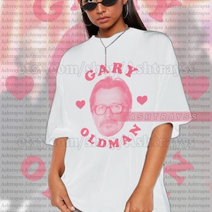 Puede incluir: Camiseta unisex blanca con un gráfico rosa de la cara de Gary Oldman, el texto "GARY OLDMAN" en rosa y corazones rosas. La camiseta es de estilo unisex.