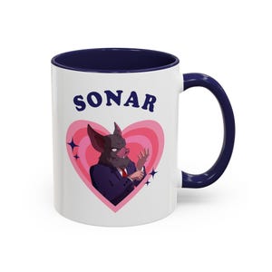 Puede incluir: Taza de cerámica blanca con interior y asa azul marino. La taza presenta una ilustración de dibujos animados de un híbrido murciélago-cerdo con traje, sobre un diseño de corazón rosa. La palabra "SONAR" está impresa encima.