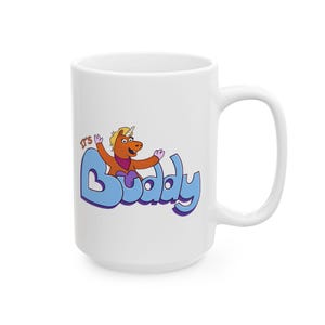 Peut inclure: Un mug en céramique blanc orné d'un personnage de licorne orange de dessin animé avec une crinière jaune et un foulard violet. Le texte "IT'S" est écrit en petites lettres orange au-dessus du grand mot bleu clair "Buddy" stylisé.