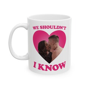 May include: White ceramic mug with a pink heart design featuring a couple kissing. The text "WE SHOULDN'T" and "I KNOW" are printed in pink on the mug. The mug has a white handle.