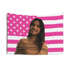 May include: A pink and white American flag tapestry with a woman in a brown off-the-shoulder top. The flag design features pink stripes and white stars. The woman is smiling and looking towards the camera.