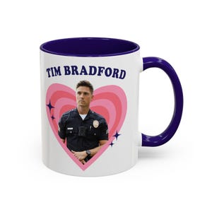 May include: White ceramic mug with a navy blue interior and handle. The mug features the text "TIM BRADFORD" above an image of a man in a police uniform inside a pink heart design. The design includes pink concentric hearts and blue star accents.
