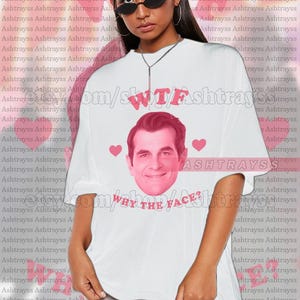 Può includere: T-shirt unisex bianca con grafica rosa raffigurante il volto di un uomo, la scritta "WTF" in rosa e la frase "WHY THE FACE?" in rosa. La maglietta ha una vestibilità ampia, adatta all'abbigliamento casual.