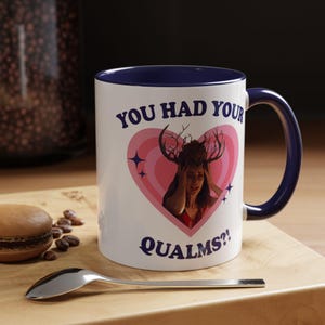 May include: A white ceramic mug with a dark blue interior and handle. The front features a pink heart graphic containing an image of a person wearing antlers, surrounded by the dark blue text "YOU HAD YOUR QUALMS?!" and small star icons.