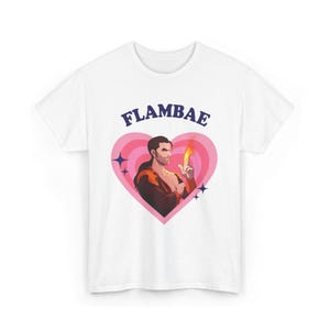 May include: White t-shirt featuring the word "FLAMBAE" above a graphic of a person holding a flame inside a pink heart. The graphic includes blue star accents.