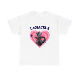 May include: A white t-shirt featuring a graphic print of a dark blue dragon creature centered inside a pink heart with concentric rings. The text LAGIACRUS is printed in dark blue capital letters above the heart, accompanied by small four-pointed star icons.