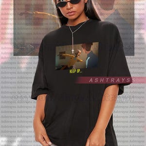 May include: Black oversized t-shirt featuring a graphic print with the text "KEEP UP..." and the brand name "ASHTRAYSS". The shirt is worn by a person wearing sunglasses.