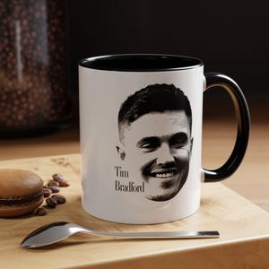 May include: A white ceramic mug with a black interior and handle, featuring a black and white portrait illustration of a person and the printed text "Tim Bradford" on the front. The mug has a smooth finish and a standard cylindrical shape.